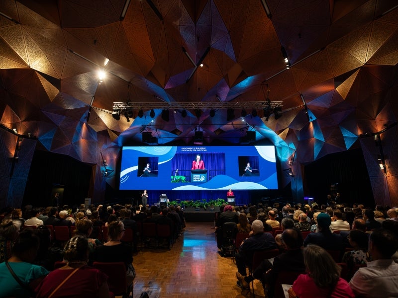 A large audience sits in a modern, geometric-ceiling auditorium watching a presenter on a brightly lit stage, with multiple screens displaying the speaker and event visuals.