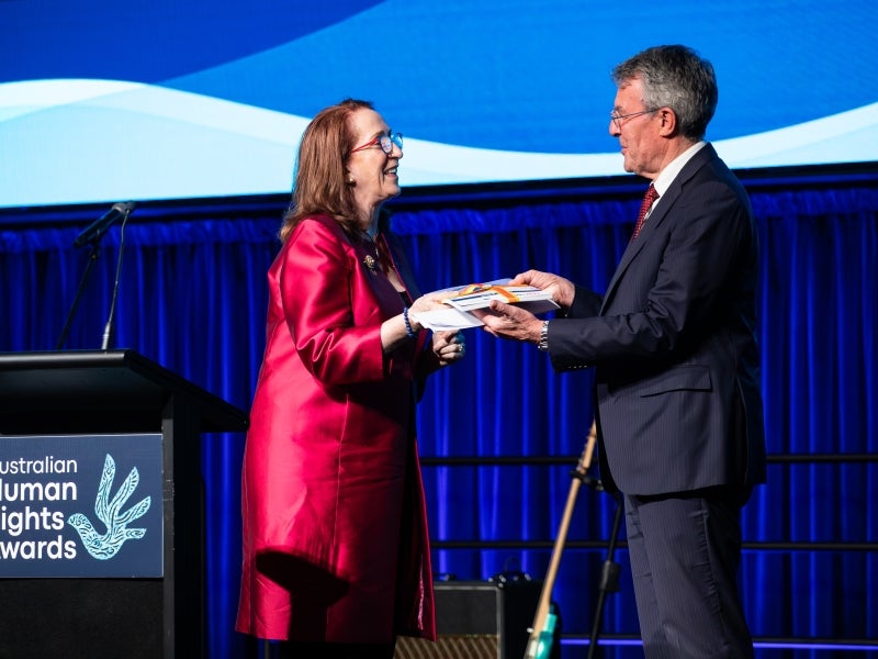 A woman in a red outfit receives an award from a man in a suit onstage at the Australian Human Rights Awards, with a podium and blue backdrop visible.