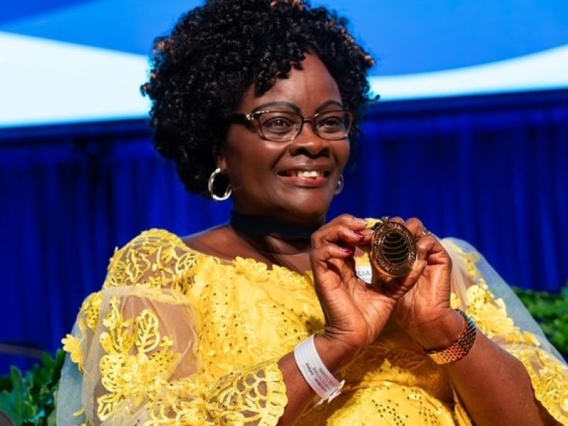 A woman in a yellow lace dress and glasses smiles while holding a spiral-shaped award. Blue stage lights and green plants are in the background.